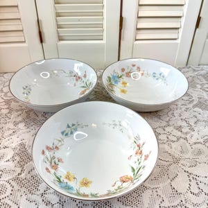 May include: Three white ceramic bowls with floral designs. Each bowl features a silver rim and a delicate pattern of colorful flowers, including yellow, blue, and orange blooms. The bowls are arranged on a white lace tablecloth.