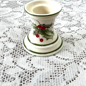 May include: A white ceramic candle holder with green trim and holly berry accents. The candle holder has a wide base and a narrow top, with a circular opening for a candle. The design includes green leaves and red berries.