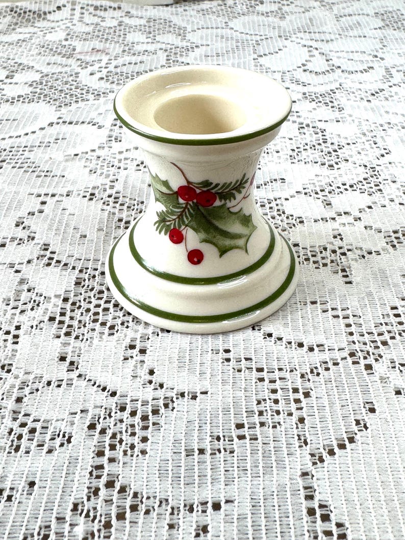 May include: A white ceramic candle holder with green trim and holly berry accents. The candle holder has a wide base and a narrow top, with a circular opening for a candle. The design includes green leaves and red berries.