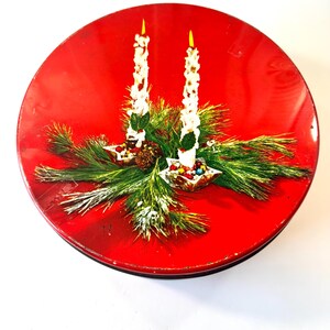 May include: A round, red tin with a Christmas-themed design. The lid features two white candles with yellow flames, surrounded by green pine needles and small, colorful candies. The tin is likely for storing cookies or other treats.