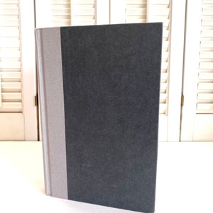 May include: A black hardcover book with a gray spine. The book is standing upright on a white surface.