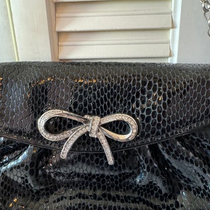 Op de afbeelding: Een zwarte clutch van nep-slangenleer met een zilveren strik versierd met strass.