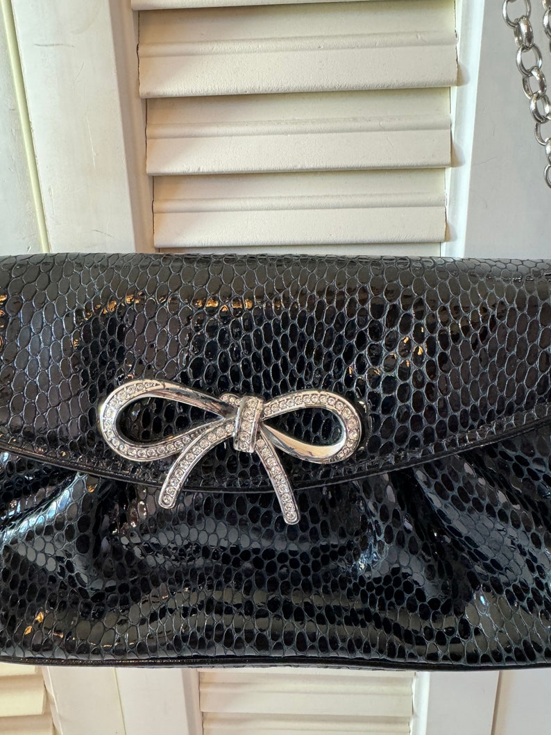 Op de afbeelding: Een zwarte clutch van nep-slangenleer met een zilveren strik versierd met strass.