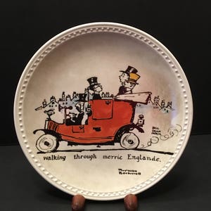 May include: A white ceramic plate with a black and white illustration of a red car with three people in it. The car is driving through a town with houses and chimneys. The text on the plate reads "walking through merrie Englande." and "Norman Rockwell".