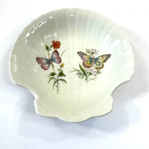 May include: A seashell-shaped dish with a cream-colored base. The dish features two butterflies and floral designs in shades of pink, yellow, and blue. The dish is approximately 6 inches wide.
