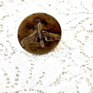 May include: A vintage, circular pin with a propeller and wing design. The pin has a bronze finish with visible wear and tear, suggesting age. The propeller is centered above the wings, creating a classic aviation emblem. The pin is likely a collectible or a piece of historical memorabilia.