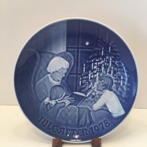 May include: Blue and white porcelain plate with a Christmas scene depicting a grandmother reading to a child by a lit Christmas tree. The plate is inscribed with "Juleaften 1978".