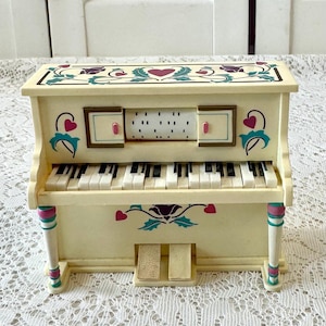 May include: A cream-colored toy piano featuring a central music roll mechanism, black and white keys, and two pedals. The exterior is decorated with painted purple and pink heart motifs, teal leaf patterns, and two vertical support columns.