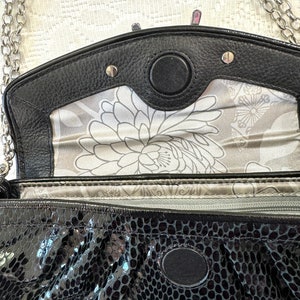 Op de afbeelding: Zwarte kettingriemtas met een slangenhuidpatroon en een zilveren kettingriem. De tas heeft een klep sluiting met een zilveren metalen accent en een bloemenvoering.