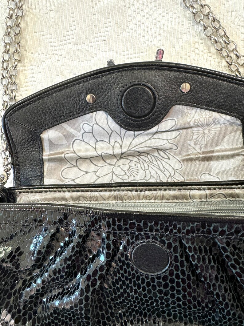 Op de afbeelding: Zwarte kettingriemtas met een slangenhuidpatroon en een zilveren kettingriem. De tas heeft een klep sluiting met een zilveren metalen accent en een bloemenvoering.