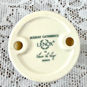 May include: Off-white ceramic decorative piece with two holes and the text "HOLIDAY GATHERINGS" and "LENOX" in green. The piece is signed "Laura Lee Luyer" and marked "&copy;LENOX".