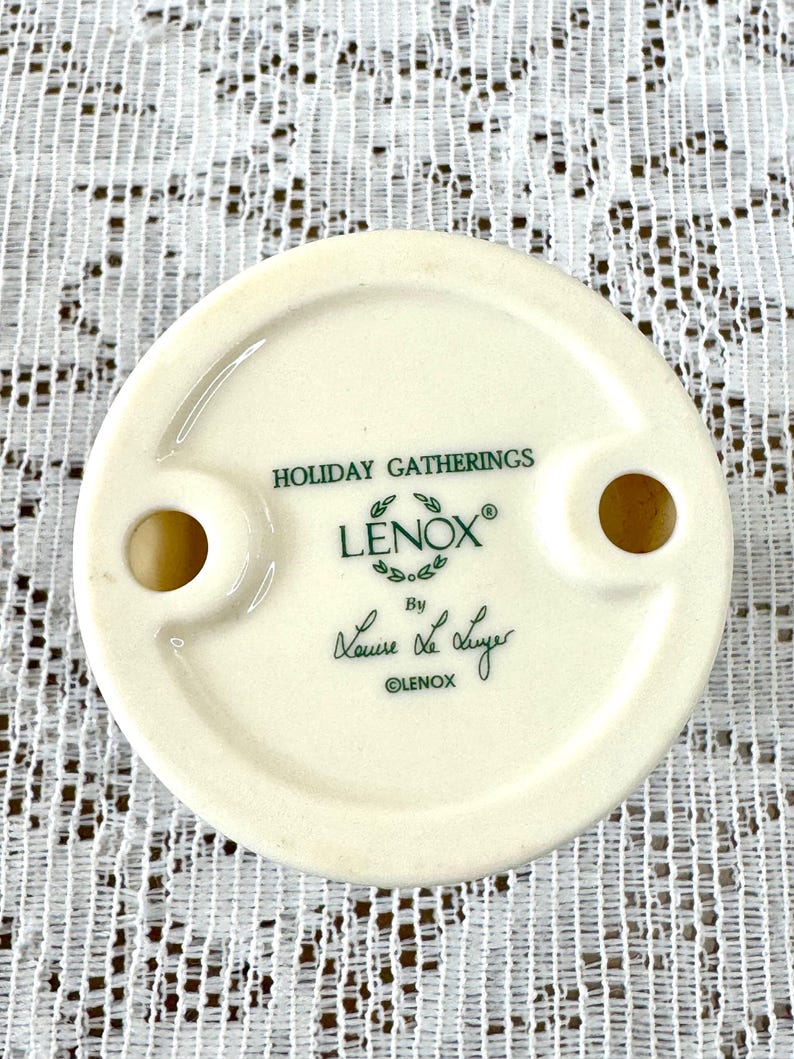 May include: Off-white ceramic decorative piece with two holes and the text "HOLIDAY GATHERINGS" and "LENOX" in green. The piece is signed "Laura Lee Luyer" and marked "&copy;LENOX".