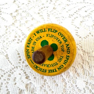 May include: A yellow wooden spinning top with a brown handle. The top features green clover-like accents and black text that reads: SPIN FAST - I WILL FLIP OVER AND SPIN ON THE STICK! - FLIPOVER TOP - MADE IN USA - THE TOYCRAFT.