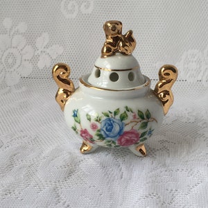 May include: A white porcelain incense burner with a floral design and gold accents. The lid features a gold figurine of a bird.