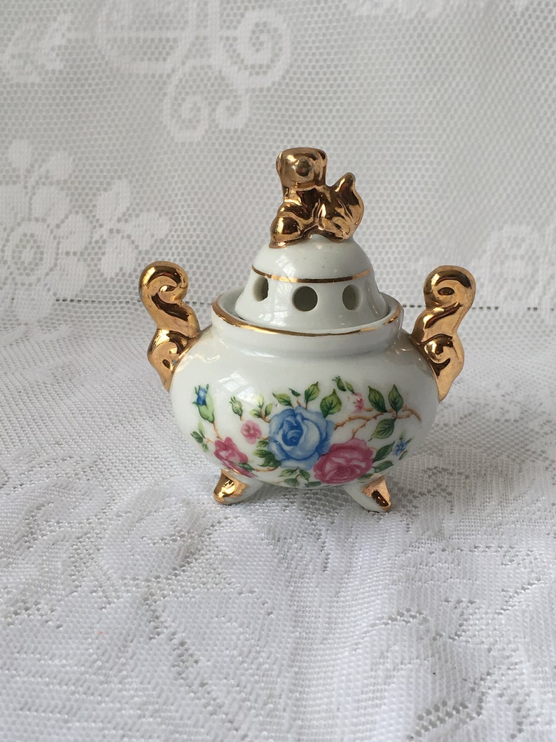 May include: A white porcelain incense burner with a floral design and gold accents. The lid features a gold figurine of a bird.