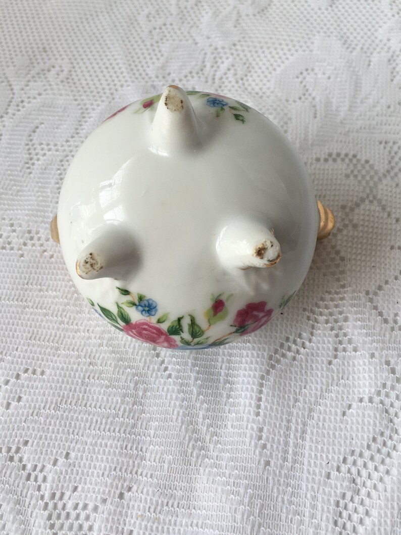 May include: White ceramic trinket dish with a floral design in pink, blue, and green. The dish has three gold feet and a rounded shape.