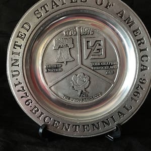 May include: A silver commemorative plate with the inscription "United States of America 1776-Bicentennial 1976". The plate is divided into four sections, each with a different image and text. The top section features the Liberty Bell and the text "1776 1976 Birth of a Nation". The right section features a stylized "YG" logo and the text "York Graphic Services, Inc. 1953". The bottom section features a white rose and the text "1741 The White Rose City".