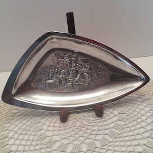 May include: A silver triangular serving tray with an embossed design of a scene with people and trees. The tray is on a wooden stand.