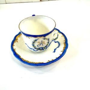 May include: A white teacup and saucer set with blue and gold trim. The cup and saucer have a scalloped edge. The cup features a blue floral design with the letters "AR". The saucer has a matching design.