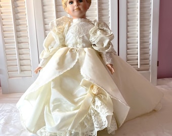 Może przedstawiać: Porcelanowa lalka ubrana w białą koronkową suknię ślubną z długim trenem. Lalka ma blond włosy ułożone w dwa kokie i niebieskie oczy.
