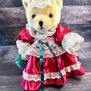 May include: A plush teddy bear wearing a red satin dress with white lace trim and a matching bonnet. The bear has a cream-colored fur and is holding a small green satin bag. The dress features a green ribbon.