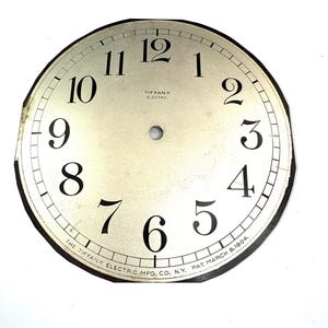 May include: An antique clock face with black numerals against a cream-colored background. The numbers 1 through 12 are displayed. The text "TIFFANY ELECTRIC" is centered, and the text "THE TIFFANY ELECTRIC MFG. CO. N.Y. PAT. MARCH 8.1904." is along the bottom edge.