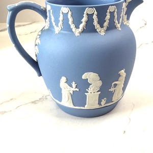 May include: A blue and white ceramic pitcher with a white floral design around the top and a white scene of three figures on the bottom.