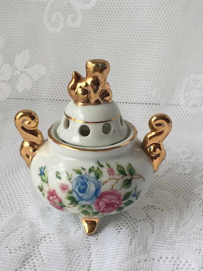 May include: A white porcelain incense burner with a gold trim and floral design. The lid features a gold figurine of a cat.