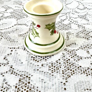 May include: Cream-colored ceramic candle holder with green trim and holly leaf and berry design. The candle holder has a wide base and a flared top, perfect for a standard-sized candle. The design is festive and suitable for the holiday season.