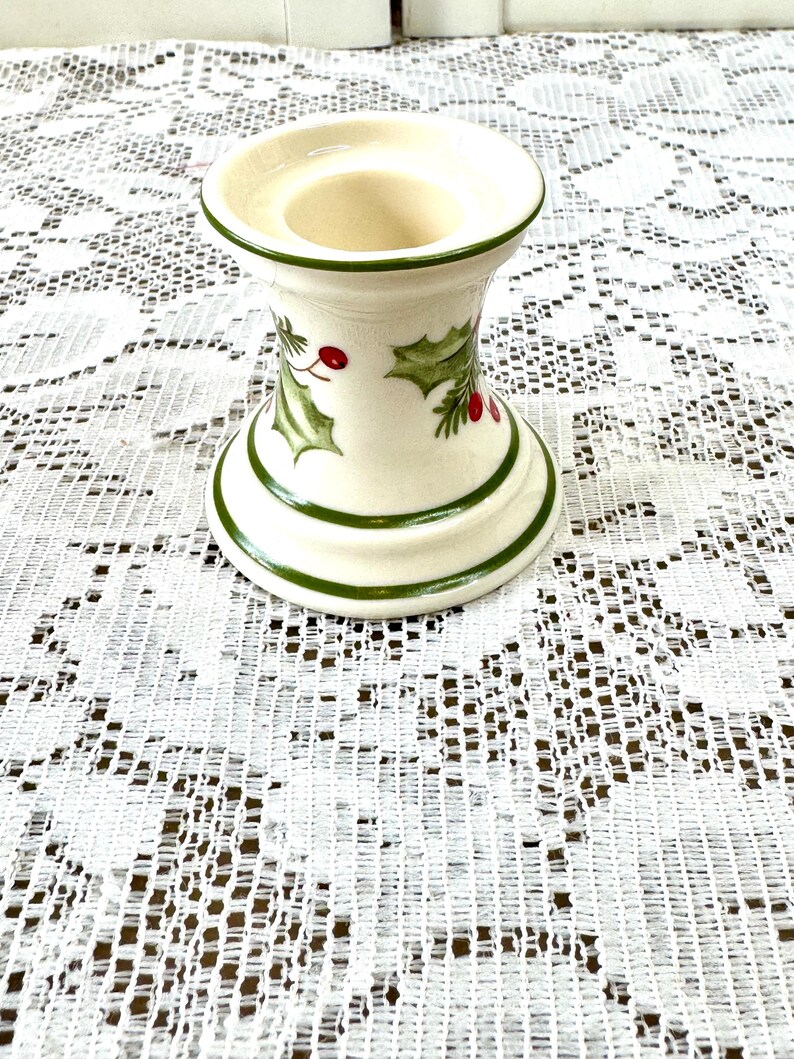 May include: Cream-colored ceramic candle holder with green trim and holly leaf and berry design. The candle holder has a wide base and a flared top, perfect for a standard-sized candle. The design is festive and suitable for the holiday season.