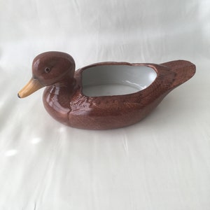 May include: A brown ceramic duck planter with a white glazed interior. The duck planter is a decorative home decor item.