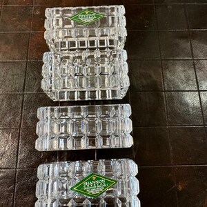 May include: Four clear, rectangular crystal ashtrays with a geometric design. Two ashtrays are stacked, and two are side-by-side. Each ashtray has a green and white label that reads "Shannon Crystal Designs of Ireland 24% Lead Crystal Hand Crafted Poland."