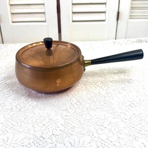 May include: A small copper saucepan with a lid and a black handle. The pan has a round body and a lid with a black knob. The handle is attached with brass hardware. The pan is sitting on a white lace tablecloth.