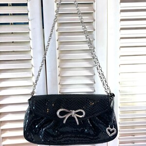 Op de afbeelding: Een zwarte leren handtas met een zilveren kettingband en een zilveren strik met strass. De tas heeft een hartvormige hanger.