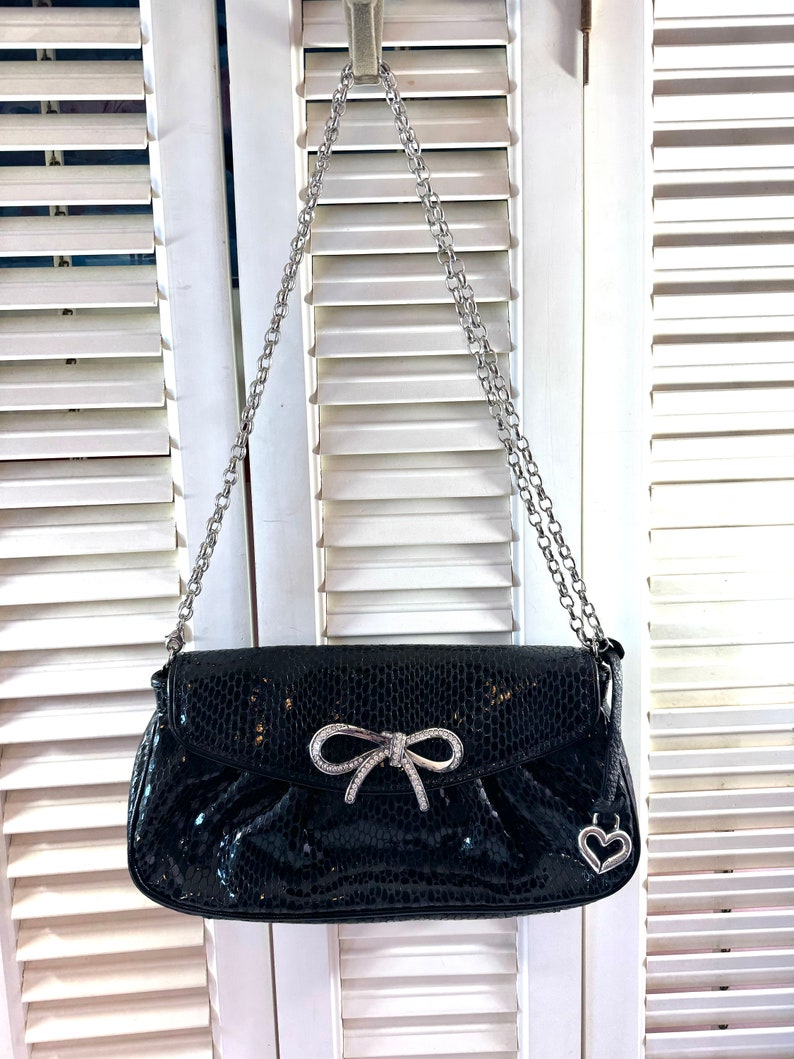 Op de afbeelding: Een zwarte leren handtas met een zilveren kettingband en een zilveren strik met strass. De tas heeft een hartvormige hanger.