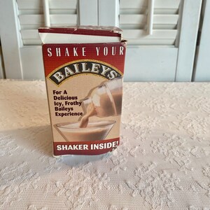 May include: A brown cardboard box with the words "Shake Your Baileys" printed on the front. The box features a picture of a glass of Baileys Irish Cream being poured from a bottle. The text "For a Delicious Icy, Frothy Baileys Experience" is printed on the box. The box also says "Shaker Inside!"