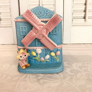 May include: A blue and pink ceramic windmill figurine with a small, pink, cartoon bunny on the bottom. The windmill has a white and pink flower design and a pink and white tulip design.