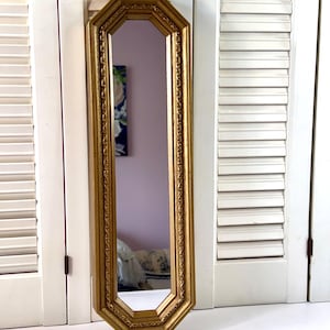May include: A tall, narrow, octagonal wall mirror featuring a metallic gold frame with a decorative, textured border. The mirror has a vertical orientation and a reflective glass surface that shows a partial view of a room interior.