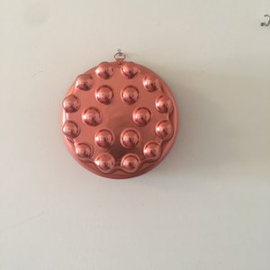 May include: A round, copper-colored kitchen mold with a series of evenly spaced, raised circular bumps. The mold is hanging on a white wall by a small metal loop. The overall shape is circular.