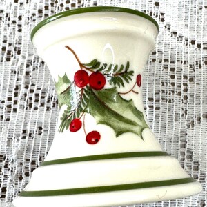 May include: A white ceramic candle holder with a green rim and base stripes. The holder features a holly and berry design in green, red, and brown, perfect for holiday decor. The candle holder is approximately 4 inches tall.