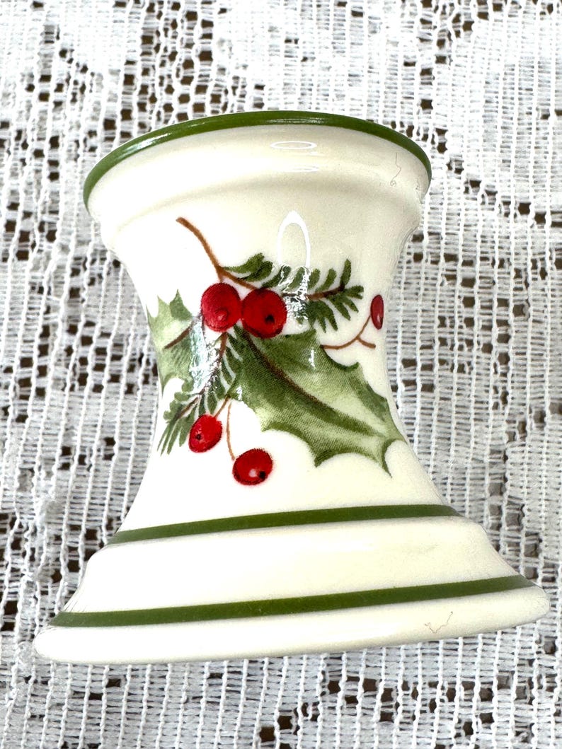 May include: A white ceramic candle holder with a green rim and base stripes. The holder features a holly and berry design in green, red, and brown, perfect for holiday decor. The candle holder is approximately 4 inches tall.