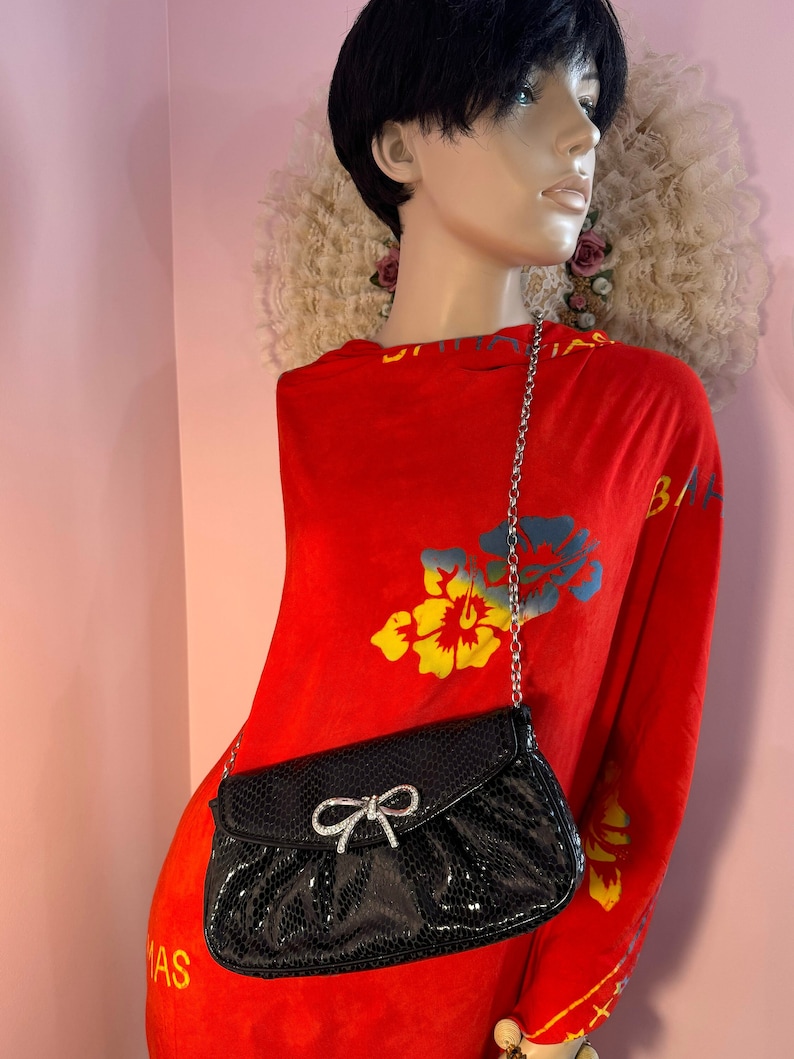 Op de afbeelding: Een zwarte clutch van imitatieleer met een strik van strass en een zilveren ketting.