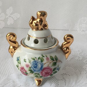 May include: A white porcelain incense burner with a gold trim and floral design. The lid features a gold figurine of a cat.