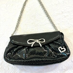 Op de afbeelding: Een zwarte handtas van imitatieleer met een zilveren kettingband, een detail van een strik met strass en een hartvormige hanger.