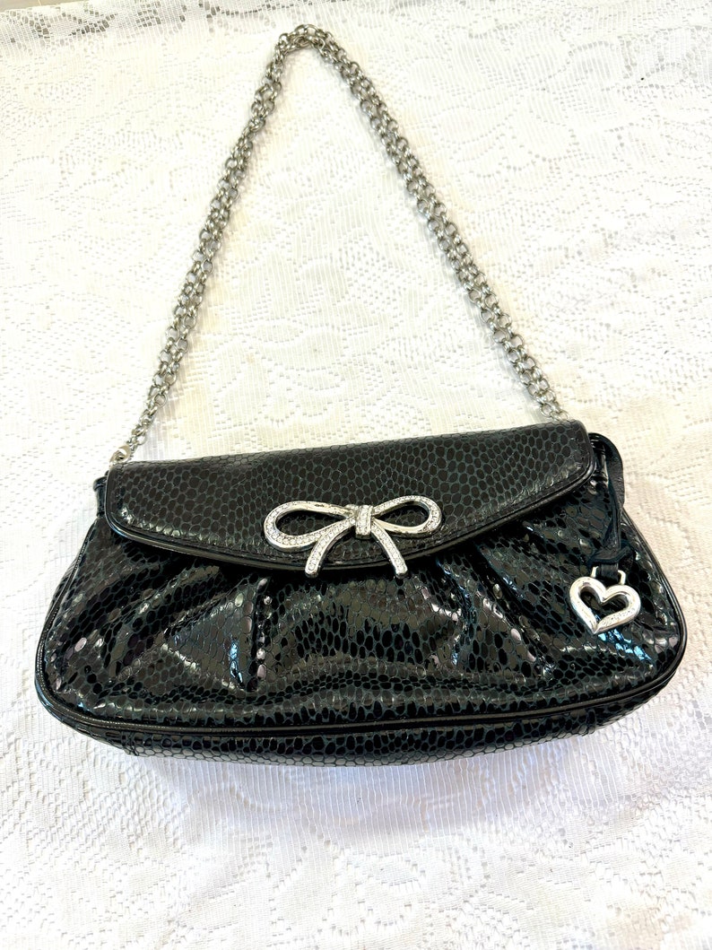 Op de afbeelding: Een zwarte handtas van imitatieleer met een zilveren kettingband, een detail van een strik met strass en een hartvormige hanger.