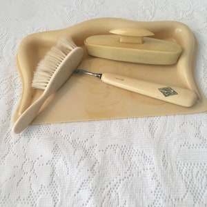 May include: A white vanity set with a brush, a small dish with a lid, and a small tool with a handle. The set is on a white lace tablecloth.