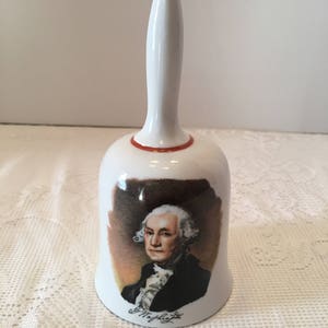 May include: White porcelain bell with a red rim and a portrait of George Washington. The bell has a white handle and the signature "G. Washington" is printed on the bottom.