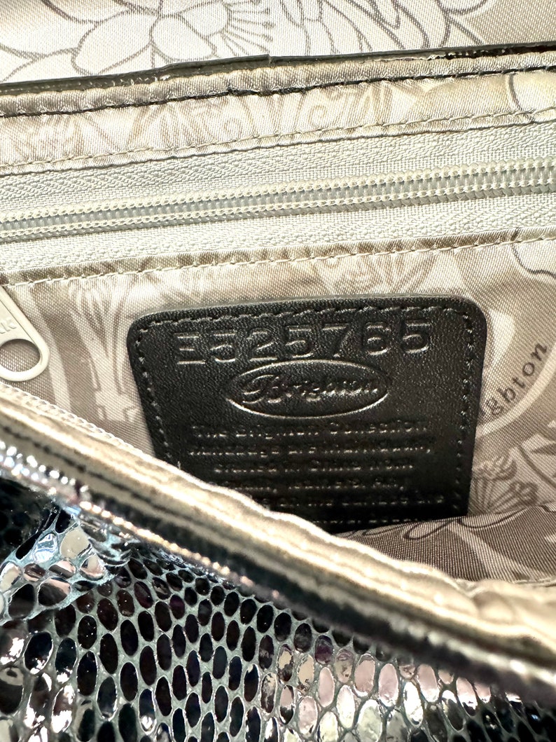 Op de afbeelding: Close-up van de binnenkant van een zilveren metallic tas met een zwart lederen label waarop "Fossil" en "E525765" staat. De voering van de tas heeft een bloemenpatroon.