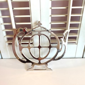 Puede incluir: Salvamanteles de plata con forma de tetera con un diseño decorativo. El salvamanteles tiene una base redonda y un asa.