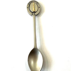 May include: A silver-colored souvenir spoon with a round emblem featuring a gold-colored lighthouse and the words "Outer Banks N.C." The spoon has a long handle and a rounded bowl.